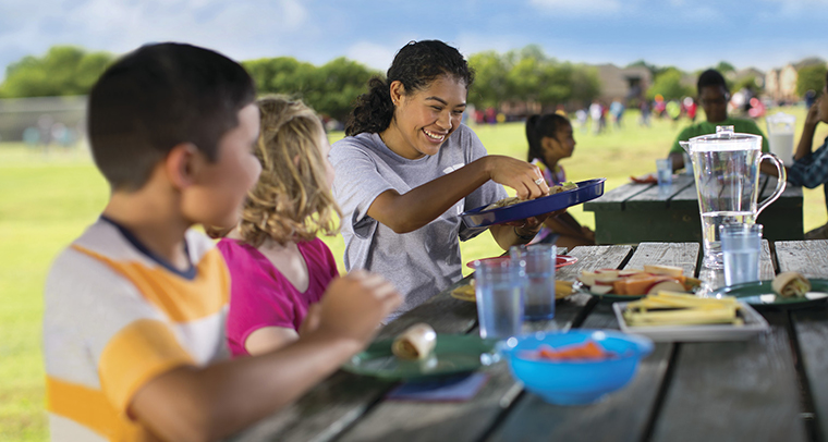 Smiling YMCA staff member and children sharing lunch at a picnic table