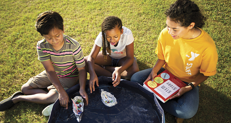 YMCA staff member sitting outside with two kids doing a science project