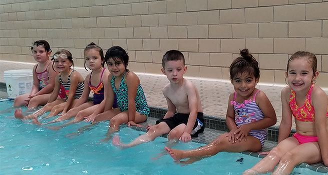 Group of kids in swimming suits sitting on the edge of an indoor pool